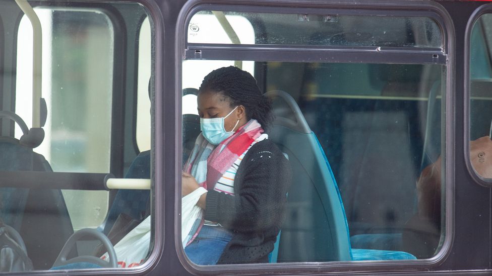 A young Black woman sits in a bus, wearing a mask.