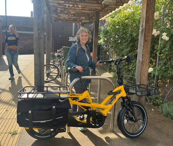 Jill Shacklock wearing denim jacket and standing behind a yellow electric cargo bike, The bike has racks at the front and back and two black paniers
