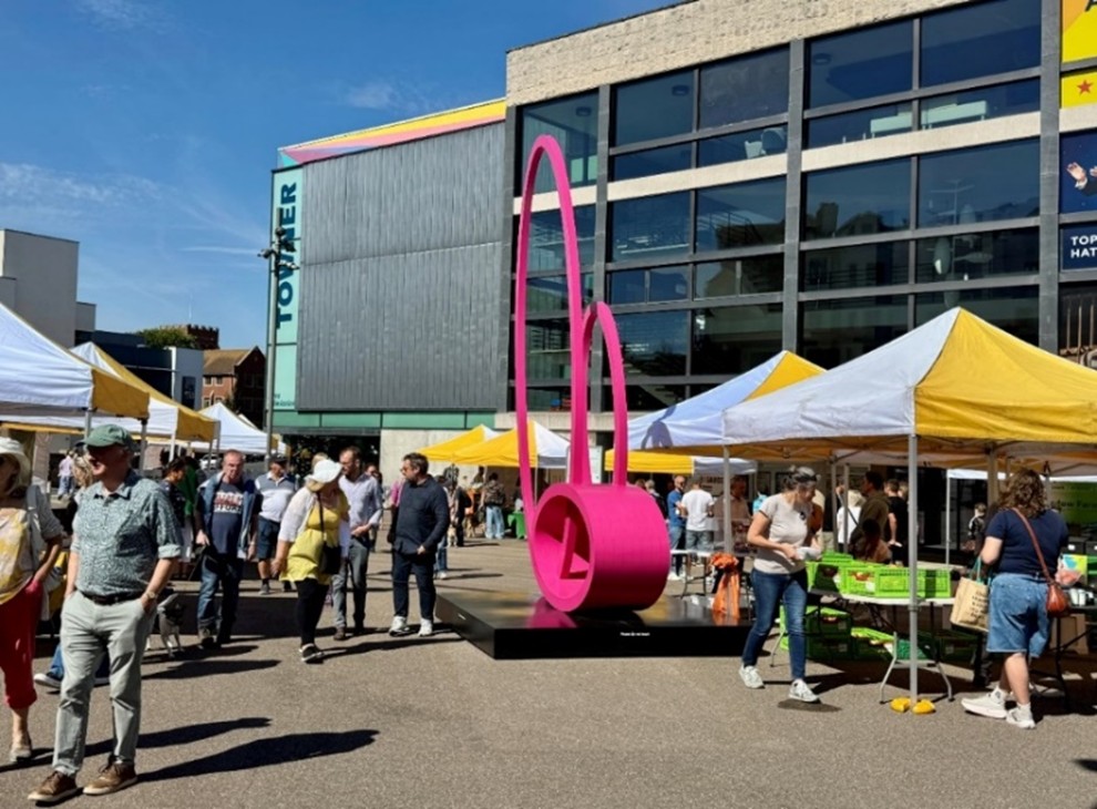 Yellow and white market stalls and a bright pink sculpture in front of the Towner gallery, creating a colourful scene full of people