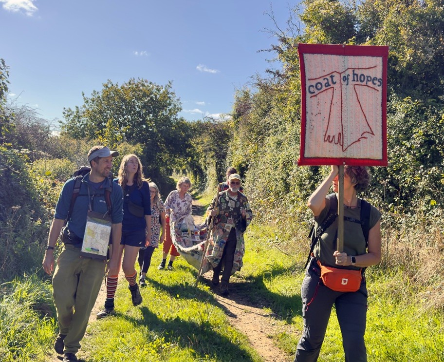 a group of people walking along countryside path, one person holds the Caot of Hopes banner, one person is wearing the coat of hopes and another person is carrying the train of the coat