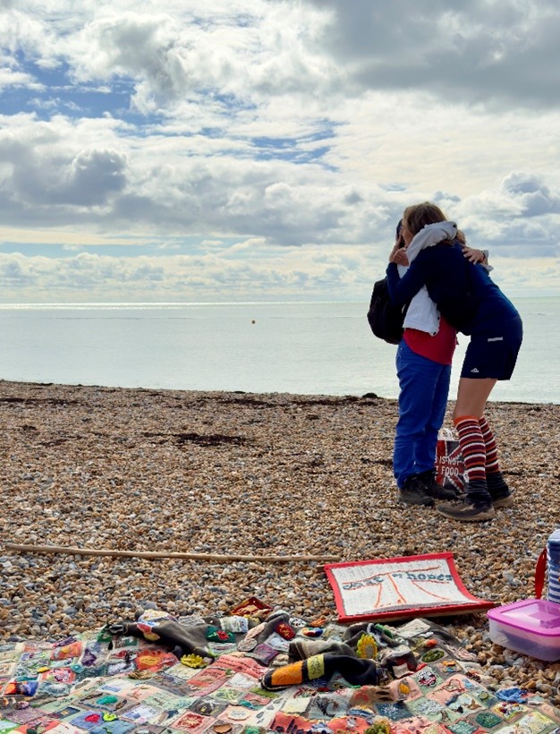 Coat of Hopes and Coat of Hopes banner lying on a pebbled beach. Two people hugging in front of the sea.