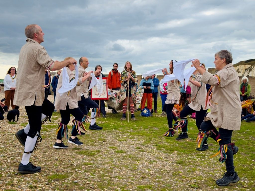 Morris dancer without hats, wearng smocks and black knee length breeches. Person wearing Coat of Hopes standing at the head of the dance formation