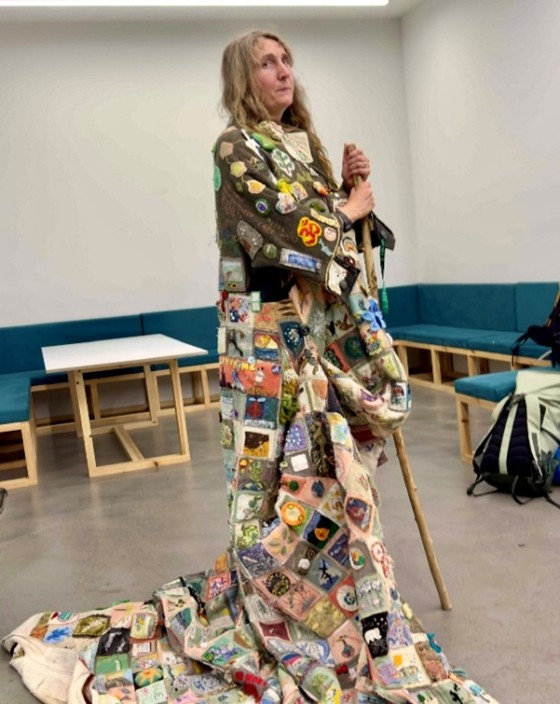 Barbara Keal with lon loose hair wearing the patchwork coat and holding a tall stick. the coat has a train which surrounds Barbara.